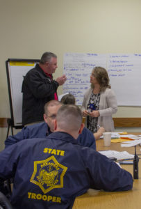 Image of a professional meeting involving Arkansas State Troopers and a civilian woman. Two male troopers, one facing the camera, are seated at a table, attentively participating in the discussion. They are wearing navy blue uniforms with 'STATE TROOPER' patches. Standing next to a whiteboard, another officer discusses a list titled 'Task Board' with the woman, indicating various action items related to a child rescue operation. The environment suggests a focus on collaborative crisis management.